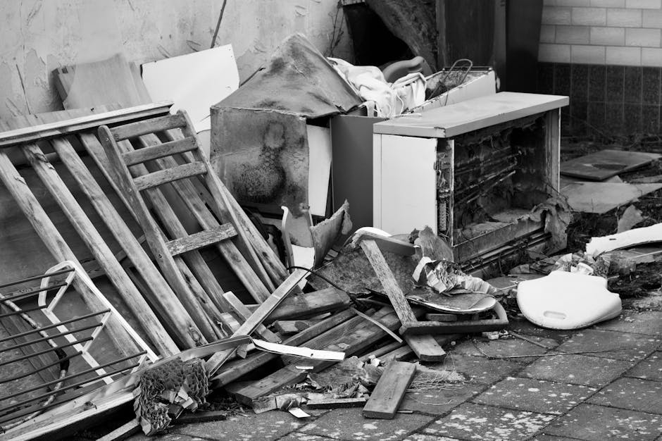 A cluttered outdoor scene featuring disorganized debris and refuse positioned against a brick wall and concrete pavement, with a broken wooden pallet leaning on the left side and various discarded ite
