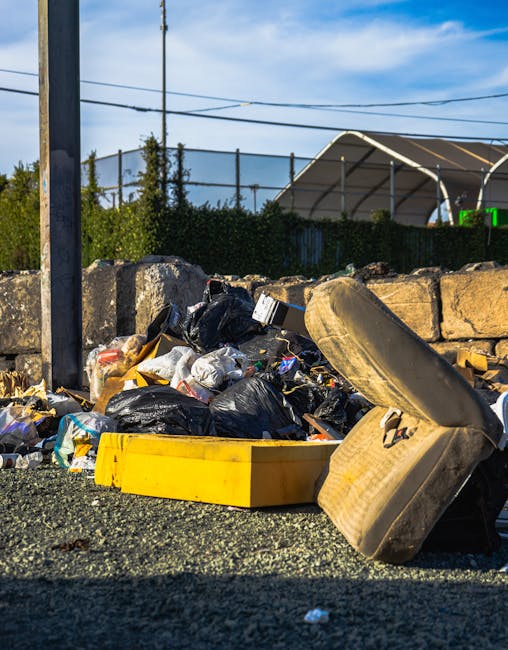 Two large black plastic garbage bags filled with waste are positioned on a paved sidewalk near a black metal fence. One bag is slightly leaning against the fence, while the other rests flat on the gro